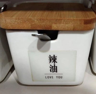 一张展示厨房用品的图片，主体是一个白色方形容器，顶部配有木质盖子。容器正面贴有标签，上方用黑色中文写着“辣油”，下方则用英文写着“LOVE YOU”。这是一个谐音梗设计，利用“辣油”(là yóu)的发音与“LOVE YOU”相似创造趣味效果。容器侧面有一个金属小勺子从开口处伸出，整体看起来像是一个实用的厨房调料罐。