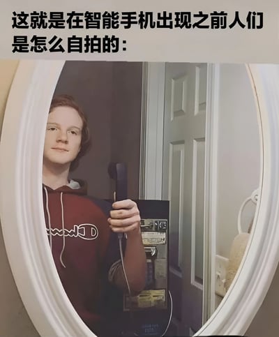 图片上方配有文字“这就是在智能手机出现之前人们是怎么自拍的：”，下方是一张真人照片。照片中一名穿着红色连帽衫的人站在镜子前，右手拿着老式座机电话的听筒举在面前，模仿现代智能手机自拍的姿势，通过镜子反射来“拍摄”自己。整体通过夸张对比的方式，幽默调侃了智能手机普及前后自拍方式的巨大差异，营造出搞笑怀旧的氛围。