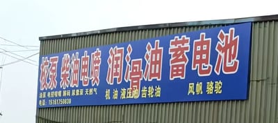 这是一张线下实拍的汽修门店户外招牌照片，招牌安装在灰绿色波纹彩钢建筑外墙上，整体为蓝底。招牌上半部分是带白色描边的红色醒目大字，内容为“校泵 柴油电喷 润滑油 蓄电池”，表明门店主营的汽修相关服务及产品；下半部分是黄色小字，左侧标注了更多服务项目：油原、电控尿素、解码、尿素泵、天然气，还有联系电话15161750838，中间标注了主营的油类产品：机油、液压油、齿轮油，右侧标注了销售的蓄电池品牌：风帆、骆驼。其中“润滑油”的“滑”字存在书写失误，字形结构异常，看起来视觉上容易引发趣味联想，这也是该图的传播亮点。