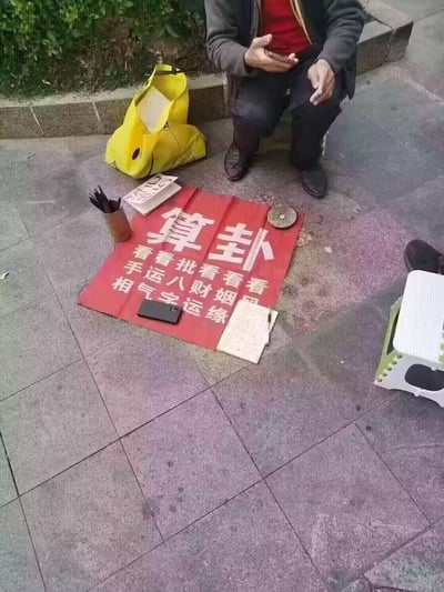 一位老人坐在街边石阶上摆摊算命，面前铺着红色布幡，上面用白色大字写着“算卦”及“看手相、看运气、批八字、看财运、看姻缘”等服务项目。布幡旁放着黄色布袋、几本书籍、笔筒和罗盘，地面为灰色石板路，背景有绿色植物，展现了传统街头算命的生活化场景。