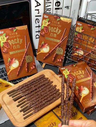 这是一张零食晒图，画面主体是格力高推出的全麦可可仁脆口味百奇饼干。画面中摆放了多盒该款百奇的产品包装盒，背景可见笔记本电脑、书籍、金属收纳篮等桌面物品，前景的浅色木质托盘上整齐摆放了十余根拆封的同款百奇饼干棒，右下角有一只手捏着三根饼干棒向镜头展示，饼干表面覆盖巧克力层，带有可可仁颗粒形成的凹凸纹理，产品包装上标注有全麦、低含糖量等相关宣传标识，整体氛围是日常桌面分享零食的场景。