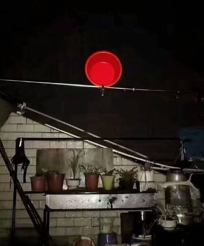 夜晚场景下，一个鲜红色的塑料盆被倒挂在一根水平的晾衣绳上，下方是一个金属架子，上面摆放着多个不同颜色和大小的花盆，里面种着植物。背景中可以看到部分砖墙和楼梯结构，整体环境看起来像是某个家庭的阳台或室外角落。光线较暗，主要依靠环境光拍摄，红色的盆在黑暗中显得格外醒目。