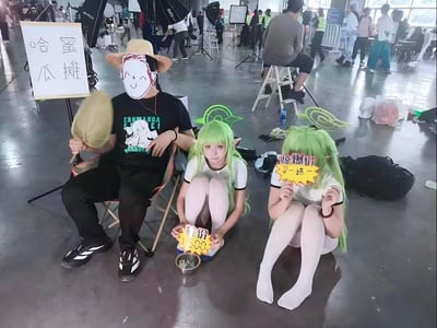 这是国内漫展现场的趣味整活场景，左侧的人戴着Q版卡通面具，坐在折叠椅上手持蒲扇，旁边立着写有「哈密瓜摊」的白色招牌；中间和右侧是两位cosplay成绿发精灵造型的人，分别举着「特价500」和「惊爆价 买一送一」的宣传牌，模仿街头卖哈密瓜的摊位。这里玩了谐音梗：绿发造型对应哈密瓜的绿色，同时暗合ACG角色C.C.的谐音，现场氛围轻松搞笑，是漫展爱好者自发的趣味互动摆拍。