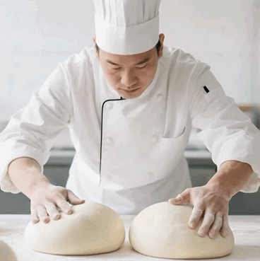 这是一段动态图（GIF），画面中一位身着全套白色厨师制服的男性厨师，正低头专注地用双手同时按压、揉制操作台上的两大团蓬松的白色面团，动作连贯自然，真实还原了面点制作过程中揉面的后厨工作场景，充满生活化的烟火气。