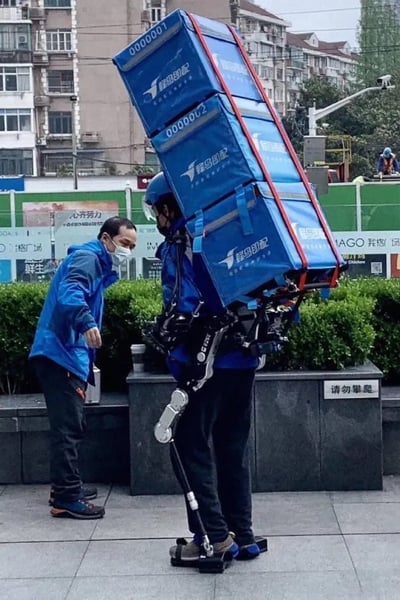 这是一张户外实拍照片，画面中一名身着蜂鸟即配蓝色工装、佩戴头盔的配送员，穿戴了机械外骨骼设备，背上用绑带堆叠固定了三个蓝色的蜂鸟即配配送箱，外骨骼的腿部支撑结构帮助承载配送箱的巨大重量，旁边另一名穿蓝色外套佩戴口罩的工作人员正低头弯腰，伸手检查调试外骨骼设备，背景是城市居民楼、施工围挡和街边绿化带，场景是物流企业对配送用外骨骼设备进行实地负载测试。