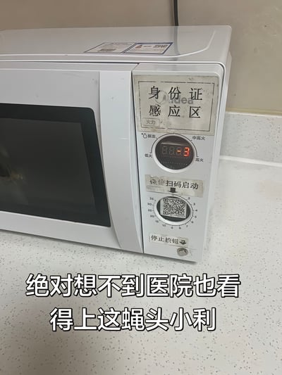 整张图展示了放置在医院公共区域台面上的一台白色美的牌微波炉，微波炉的操作面板被人为张贴了多个标识：原本的火力调节旋钮上方贴有写着“身份证感应区”的贴纸，时间调节旋钮上方贴有“微信扫码启动”的贴纸并覆盖了一个二维码，旋钮下方的小按钮旁贴有“停止按钮”的贴纸，原本的微波炉调节功能被改造，用户需要扫码或刷身份证才能启动使用。图片底部叠加了白色黑边的吐槽文字“绝对想不到医院也看得上这蝇头小利”，直白表达了拍摄者对医院将公用微波炉改为收费使用、赚取小额收益的不满情绪。