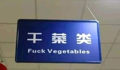这是一张悬挂的蓝色品类标识牌的实拍图，标识牌上方为白色简体中文“干菜类”，是用于指示商超、菜市场等场所的干菜区域的标牌，但下方的英文翻译却错误地使用了带有冒犯性的“Fuck Vegetables”，正确的干菜类英文翻译应为“Dried Vegetables”，这种明显的翻译失误让原本普通的标识牌产生了滑稽恶搞的效果，成为网络上的搞笑素材。