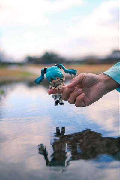 这是一张充满创意的手办摄影作品，利用透视构图与水面反射的视觉效果，将GSC出品的初音未来粘土人手办放置在人的指尖上，营造出玩偶仿佛正悬停在水面上方、即将触碰水面的童话场景。水面清晰倒映出手办的身影，背景是模糊的户外水域与远处的朦胧景物，整体氛围治愈温馨，没有双关、谐音梗或暗示内容。