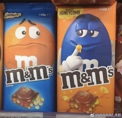 图片展示了两款M&M's巧克力的包装对比：左侧为蓝色背景的CRISPY口味，包装上的橙色M&M's角色眉头紧锁、嘴角下撇，呈现出愁眉苦脸的表情；右侧为橙色背景的HONEYCOMB口味，蓝色M&M's角色则眼神得意、嘴角上扬，并用手指着嘴边的蜂巢脆片，表情自信又带点小调皮。两款包装的角色表情形成鲜明反差，营造出幽默有趣的视觉效果。