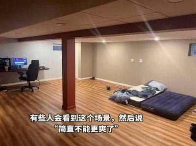这是一张静态实拍图，展示了改造后的地下室私密空间：房间铺设浅棕色木质地板，中间有一根红棕色承重立柱将空间大致分隔，墙面简洁干净。左侧区域布置了电脑桌，桌上摆放着处于开机状态的显示器，桌前搭配一把黑色电竞椅，作为娱乐办公区域；右侧靠墙位置放置了床垫和充气床，上面堆放着被褥枕头，作为休息区域。图片底部配有中文字幕，内容为“有些人会看到这个场景，然后说“简直不能更爽了””，表达了部分人群对这种可以就近兼顾娱乐、工作和休息的独立私密空间的喜爱与向往。