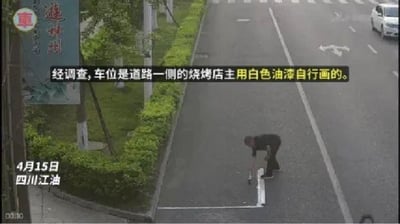 这是一张监控画面截图，拍摄时间为4月15日，地点为四川江油。画面中可见一名男子在靠近绿化带的道路边缘弯腰用白色油漆绘制停车位标线，画面配有字幕说明经调查这些车位是道路一侧的烧烤店主自行用白色油漆绘制的，属于违规行为，画面远处还有正常行驶的白色车辆，道路旁有整齐的绿化植被和标识牌。