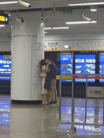 广州地铁天河公园站的站台上，一对情侣依偎在立柱旁相拥亲吻。背景可见地铁列车门和蓝色电子屏，屏幕上滚动显示着“来已来 共赴征”等文字及部分ETF信息，站台地面光洁，顶部有监控摄像头和照明设备，整体呈现出都市通勤中的温馨瞬间。