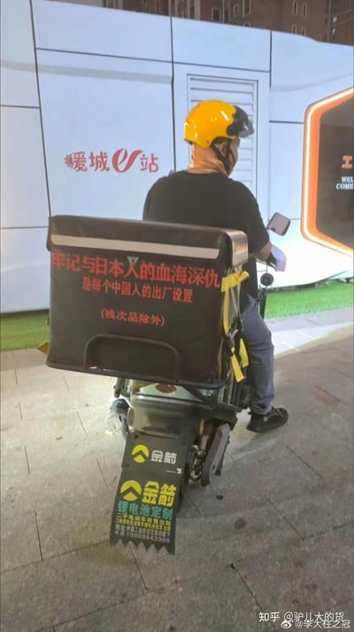 图片展示了一名外卖骑手的背影，他戴着黄色头盔，骑着印有'金箭'品牌的电动车。骑手的配送箱上贴有红色争议标语：'牢记与日本人的血海深仇 是每个中国人的出厂设置 (残次品除外)'。背景中可见'爱城U站'的白色建筑。