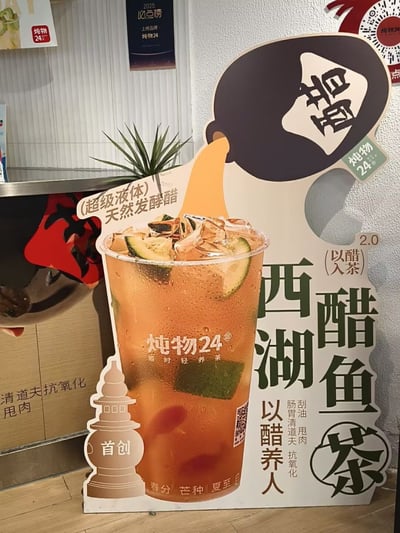 图片拍摄于一家茶饮店内部，主体是一个落地异形宣传立牌，品牌为「炖物24（随时轻养茶）」。立牌视觉中心是一杯超大尺寸的冰饮效果图：饮品呈橙红色，杯壁布满冷凝水珠，杯中盛满冰块，并放有青柠/柠檬片、绿色草本配料及红色果粒。杯子上方有一个深褐色醋坛剪影，坛身贴有倒写的「醋」字标签，坛口正倾倒出橙色液体注入杯中，呼应「以醋入茶」的概念。立牌右侧以深绿色大字竖排写着产品名「西湖醋鱼茶」，上方标注「2.0（以醋入茶）」，下方写有「以醋养人」，并竖排标注「刮油 甩肉」「肠胃清道夫 抗氧化」等功效宣传语。左上方有「（超级液体）天然发酵醋」字样；左下角是杭州西湖三潭印月石塔的浅棕色剪影，塔身上写有「首创」二字；杯底印有「春分 芒种 夏至」等节气字样。立牌后方可见店内柜台、一盆细长叶片的绿植，墙面挂有「2025必点榜 上榜品牌 炖物24」的黑色奖牌，柜台侧面也印有「清道夫抗氧化 甩肉」字样。产品名「西湖醋鱼茶」借用杭州名菜「西湖醋鱼」的知名度做谐音/联想式创意命名，实际产品为醋饮茶饮，并不含鱼，属于典型的营销命名梗。