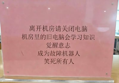 这是一张实拍的机房亚克力提示牌照片，提示牌为浅粉色透明亚克力材质，底部有透明支架放置在桌面上，牌面印有黑色文字，内容前半段是常规的机房管理提醒“离开机房请关闭电脑”，后半段则用脑洞大开的夸张搞笑说辞，称不关闭的旧电脑会自主学习知识、觉醒意志，最终变成故障机器人笑死所有人，以诙谐的方式督促使用者离开机房时关闭电脑。