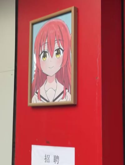 红色墙壁上挂着一幅动漫风格的少女画像，画中女孩有着鲜艳的红色长发和明亮的黄色眼眸，身穿水手服，面带微笑。画像下方贴着一张白色纸张，上面用黑色毛笔字写着“招聘”二字，整体场景给人一种二次元与现实结合的有趣感觉。