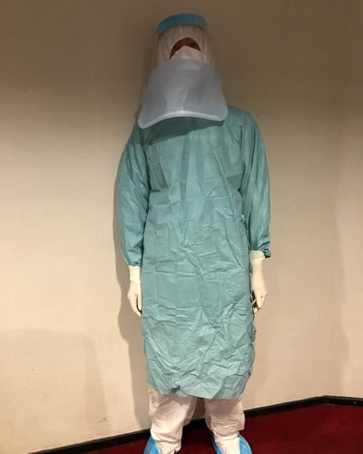 图片中展示了一位身着全套医疗防护装备的人员，包括青色防护服、白色手套、蓝色鞋套和透明面罩，站立在浅色墙壁和红色地毯的室内环境中。