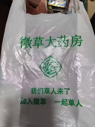 这是一张微草大药房的白色塑料购物袋实拍图，袋身采用绿色油墨印刷品牌相关内容。袋身最上方是品牌名称“微草大药房”，中间位置是带有草类植物图案的品牌专属标识，下方依次印有宣传语“我们草人来了”“加入微草 一起草人”。其中“草人”是谐音“超人”的趣味用法，属于谐音梗营销，通过这种接地气又有记忆点的方式，打造品牌专属的宣传话术，增强宣传的趣味性和传播性。