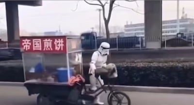 这张图片呈现了一个充满反差感的趣味场景：原本是《星球大战》里银河帝国的暴风兵（白兵），却以cos装扮出现在现实城市道路上，骑着一辆搭载煎饼摊的三轮车行进。摊位上方的红色招牌写着“帝国煎饼”四个字，巧妙结合了暴风兵所属的“帝国”设定，将科幻IP元素与接地气的街头美食摊点结合，制造出强烈的违和感与搞笑效果，属于网友的创意cos摆拍内容。背景能看到城市的道路、绿化带和远处的建筑物，是日常的城市街景。