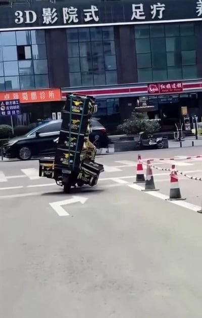 一张城市街道实景照片，画面中央一辆美团外卖三轮车疑似发生侧翻事故，车身倾斜严重，车上的外卖箱摇摇欲坠。背景可见多家商铺招牌，包括"3D影院式足疗"、"皇雅岚诊所"和"红娘连锁"等。路面上摆放着几个交通锥和警示带，似乎是为了提醒过往车辆注意安全。整个场景给人一种意外突发的惊险感觉，展现了外卖配送工作中可能遇到的交通安全风险。