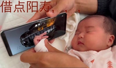 这是一张利用视觉错位效果打造的趣味梗图，画面中一名熟睡的婴儿躺在床上，家长将手机屏幕显示的游戏背包界面放在婴儿小手上方，通过拍摄角度让画面看起来像是婴儿的手伸进了游戏背包里。图片上方配有文字“借点阳寿，，，”，玩了生存类游戏圈的经典梗——玩家常调侃自己“阳寿不足”，需要“借阳寿”来获取游戏里的好运或稀缺资源，结合错位画面，营造出婴儿要从游戏背包里“借阳寿”的荒诞搞笑效果，用亲子日常结合网络热梗，充满反差感与趣味性。
