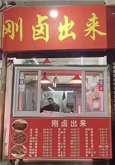 这是一张实拍的卤味小店照片，店铺门头采用红色底色配黄色大字，文字内容为“刚卤出来”，本意是宣传店内卤味为新鲜出炉的状态，但由于字体和排版设计的原因，“刚”和“卤”两个字很容易被误看成不雅脏话的谐音，产生了搞笑的歧义效果。店铺下方的红色价目表顶部同样印有“刚卤出来”字样，还标注了各类卤味的名称和对应售价，窗口内可见一名男性工作人员正在值守。