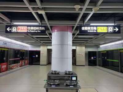这是武汉地铁中南路站的换乘站台实拍画面，画面中央的立柱分隔开两条线路的乘车区域：左侧为2号线往佛祖岭方向的站台，屏蔽门带有粉色装饰条，站台标识牌为粉色系；右侧为4号线往柏林方向的站台，屏蔽门带有绿色装饰条，标识牌为绿色系。站台中间设置了一台四盆式公共直饮水设备，整个站台空旷无乘客，顶部的线路方向指示牌清晰醒目，站点环境整洁明亮。
无谐音梗、双关或暗示内容。