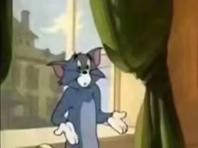 这是一张经典动画《猫和老鼠》的静态截图，画面中是主角之一的汤姆猫，它站在室内窗边，背景能看到窗外的建筑。汤姆猫眉头微皱，眼睛睁大，露出无奈又不知所措的神情，同时双手摊开，这个姿态生动地传递出无语、对现状无可奈何的情绪，常被用作表达此类情绪的表情包素材。