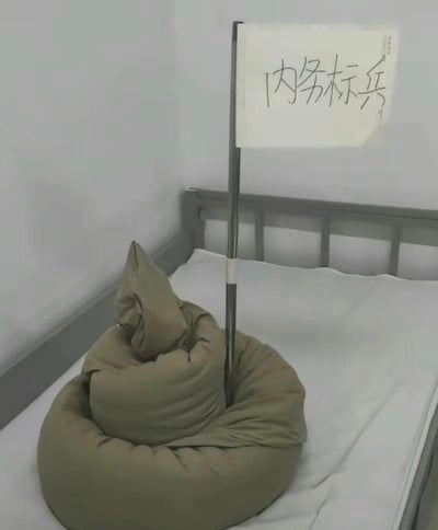图片呈现的是宿舍床铺场景，原本要求叠得方正整齐的军绿色被子，被创意性地叠成了类似马桶圈加立柱的搞怪造型，立柱上还插着一张手写“内务标兵”的白纸。这种造型和标准内务要求形成强烈反差，用反讽的方式调侃严格的内务整理规则，充满戏谑搞笑的氛围。