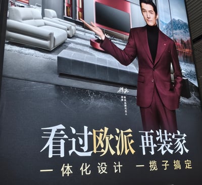 这是一张欧派家居的广告宣传图，画面主体为穿着酒红色西装的代言人胡夏，他站在一个现代风格的客厅场景前，右手伸出做出展示姿态。背景中可以看到灰色的沙发组合、红色的电视背景墙和窗外的山水景色。图片下方有醒目的广告语“看过欧派 再装家”，以及副标题“一体化设计 一揽子搞定”，突出品牌的整体家居解决方案优势。