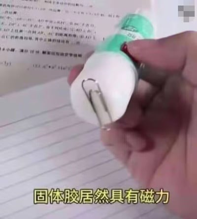 整张图为实拍画面，一只手拿着绿白外观的固体胶，固体胶的底端粘有一枚回形针，看起来仿佛是固体胶带有磁力吸住了回形针，画面底部配有黄边黑字的醒目字幕“固体胶居然具有磁力”，背景是带有打印文字和横线的纸张，实际是利用固体胶的粘性粘住回形针制造的视觉错觉，用来制造搞笑反差效果。