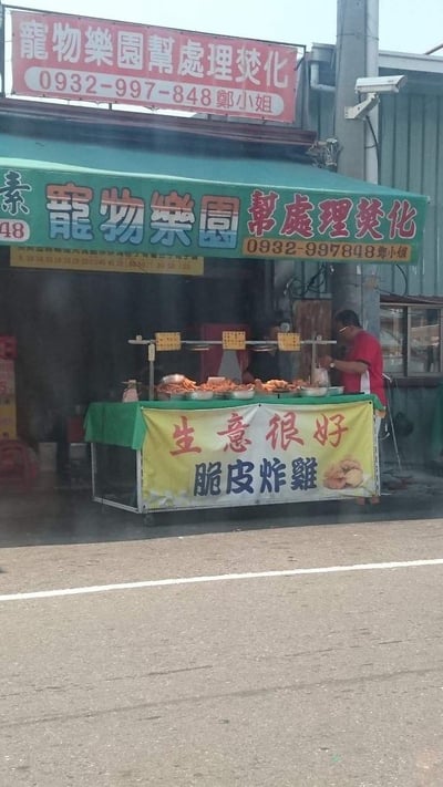 这是一张台湾地区的路边实拍图，画面里是一个售卖脆皮炸鸡的路边小吃摊，摊主正在摊位前忙碌，摊位横幅写着「生意很好 脆皮炸雞」，摆放着各类炸鸡食材。但摊位上方的遮阳篷和红色招牌却是「寵物樂園幫處理焚化」的宠物殡葬服务广告，还附带了联系人郑小姐的电话。这种完全不相关的业务出现在同一个场景里，形成了极强的荒诞反差感，让人觉得滑稽搞笑。

反差点：小吃摊的炸鸡生意和殡葬服务的招牌完全错位，形成了诡异又搞笑的视觉冲突。