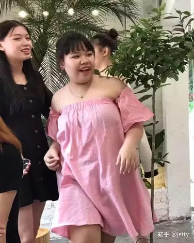 画面中心是一位穿着粉色露肩连衣裙的女生，她嘴巴大张，脸上洋溢着开心兴奋的笑容，正迈步向前，情绪十分高涨。她左侧的女生穿着黑色连衣裙，同样面带笑容看向一旁，背景中还有其他人物和绿植，场景看起来是在半户外的休闲场所，整体氛围轻松愉快，充满生活气息。