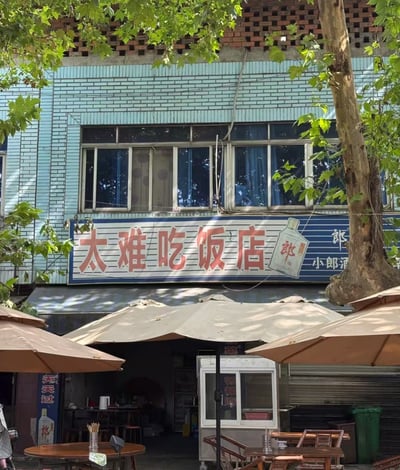 照片展示了一家位于蓝色砖墙建筑底层的餐馆，招牌醒目地写着红色大字‘太难吃饭店’，右侧配有‘小郎酒’的产品图片和文字。建筑上方是红砖结构，周围有茂密的绿色树叶遮挡部分视线。餐馆门口摆放着几张木质桌椅，上方覆盖着米色遮阳伞，营造出街边小店的氛围。最引人注目的是餐馆名称的反常规命名——‘太难吃’本是负面评价，却被用作店名，形成强烈的反差幽默，让人会心一笑。