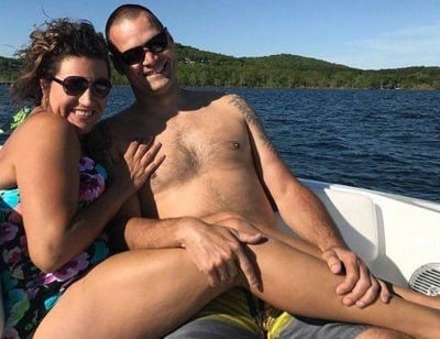 这是一张在湖面游船上拍摄的趣味真人照片，图中一男一女亲密依偎，两人都戴着墨镜，面带开心的笑容。由于女性的腿部姿势与男性的手臂位置巧合重叠，形成了视觉错位效果，看起来仿佛男性拥有女性的腿部，制造出滑稽的错觉。背景是开阔的蓝色湖水与远处的绿色山林，整体氛围轻松愉快。