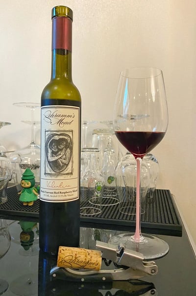 一张展示蜂蜜酒品鉴场景的照片。画面中央是一瓶Schramm's Mead Valentine系列黑加仑红莓蜂蜜酒，瓶身上有两个人物相拥的素描图案。酒瓶旁边是一个装有深红色液体的高脚杯，杯脚为粉色。前景中有一个带有品牌标识的软木塞和金属开瓶器。背景中可以看到多个不同形状的玻璃杯整齐排列在黑色托盘上，左侧还有一个绿色的小雕像装饰。整个场景呈现出精致的饮酒氛围。