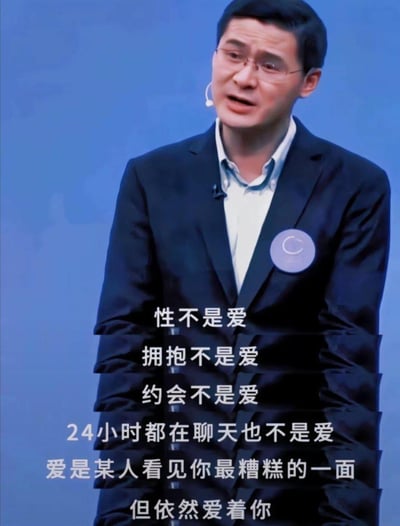 图片中是一位穿着深色西装、佩戴眼镜的男性演讲者，他站在蓝色背景前，戴着耳麦，胸前有一个圆形徽章。演讲者表情严肃认真，似乎正在发表关于爱情的观点。图片下方叠加了白色文字：'性不是爱 拥抱不是爱 约会不是爱 24小时都在聊天也不是爱 爱是某人看见你最糟糕的一面 但依然爱着你'，表达了对爱的独特定义。