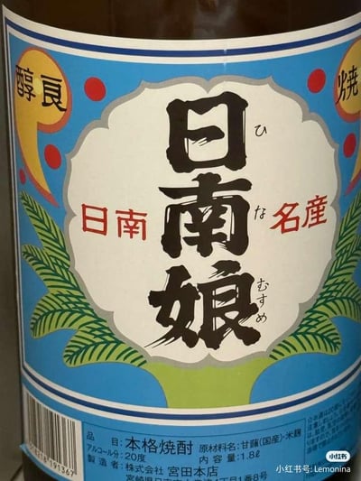 这是一张日本株式会社宫田本店出品的「日南娘」本格烧酎的商品标签实拍图。标签设计色彩明快，以蓝色为底色，搭配绿色植物纹样与红色圆点装饰，视觉风格充满地域特色。标签中央是醒目的毛笔书法字体「日南娘」，旁注日文假名「ひなむすめ」，两侧配有「日南 名産」字样，左上角标注「醇良」，右上角标注「烧」字，点明这是一款优质烧酎。标签下方的蓝色区域详细列出了商品信息：品目为本格烧酎，原材料为国产甘薯与米麴，酒精度数20度，容量1.8升，制造者为株式会社宫田本店，地址位于宫崎县日南市。