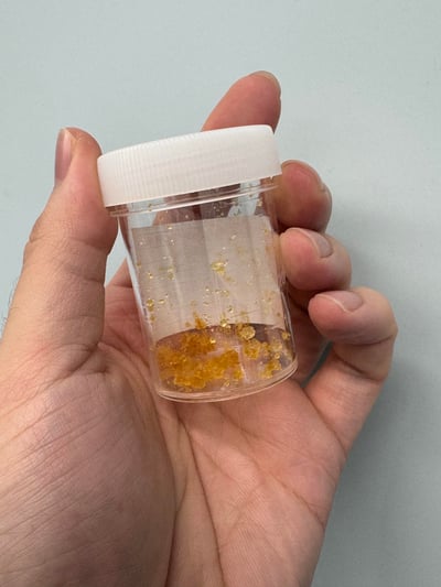 一只手掌托着一个带白色旋盖的透明小容器，容器底部和内壁附着着一些金棕色的细碎颗粒，看起来像是提炼后的金屑或金箔碎，背景是纯色浅灰墙面，画面聚焦于容器和手中的物质，整体风格平实，是对私藏材料的记录展示。