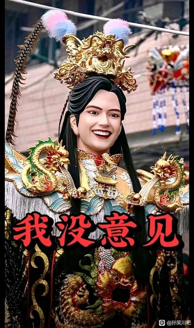 图片中展示了一位身着华丽传统服饰的人物，头戴金色龙纹头饰，装饰有粉色和蓝色绒球以及羽毛。人物面带微笑，背景中隐约可见传统节庆装饰。图片底部有醒目的红色文字'我没意见'，表达一种中立或顺从的态度。这种将传统服饰元素与现代网络用语结合的方式，形成了幽默的反差效果。