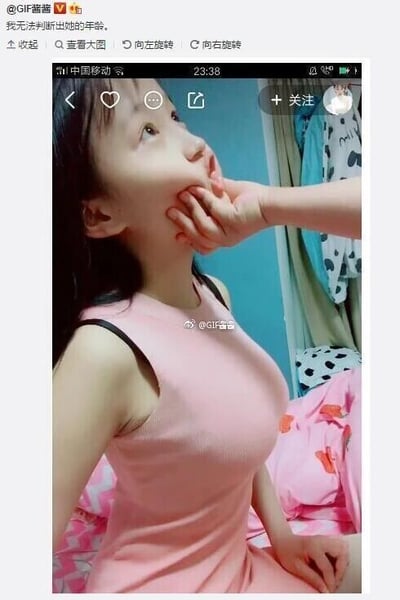 这是一张来自微博的社交媒体截图，截图内的主体是一位穿着粉色无袖上衣的女生，她的脸部被一只手捏住向上抬起，脸部神态看起来十分幼态，但身材却显得比较成熟，这种强烈的外观反差，让发布者配文表示“我无法判断出她的年龄”。背景是普通的室内卧室环境，能看到带有图案的床铺和挂着的衣物，图片中部偏下还有@GIF酱酱的水印。