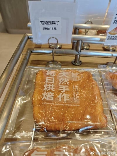 图片展示了面包店的陈列架，其中一个透明包装的烘焙食品格外显眼。上方的价格牌写着“可颂压扁了 原价:18元”，下方包装袋上印有“每日烘焙 天然手作”和“酥后 SO FULL DAILY BAKING WORKSHOP”字样。被“压扁”的可颂表面撒有芝麻，造型独特。店家通过幽默的商品命名“可颂压扁了”替代常规名称，既直白描述了商品形态，又带有调侃意味，容易引发顾客会心一笑。