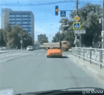 这是一段动态GIF，从车内第一视角拍摄城市道路场景，画面中看似有一辆橙色跑车在前方道路行驶，实则是后方卡车上装载的橙色货物，因视角巧合形成了“跑车在路上行驶”的视觉错位错觉。道路旁有城市建筑、交通标识，还有其他正常行驶的车辆，整体是日常的城市街景，这种错位效果让人感到新奇又搞笑。