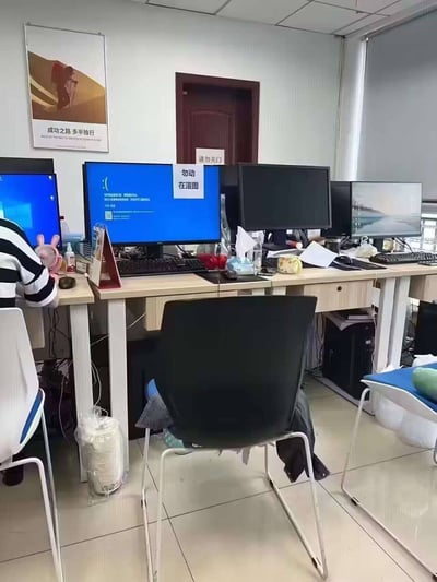 办公室场景展示，多张电脑显示器排列在办公桌上，其中一台屏幕显示蓝色界面并标注'勿动 在渲图'。一名穿着黑白条纹上衣的人正在电脑前工作，桌上有各种办公用品。墙上挂着一幅写有'成功之路多独行'的励志海报，门上贴有'请勿关门'的提示。整个场景呈现典型的办公环境，显示有人正在进行电脑渲染工作。