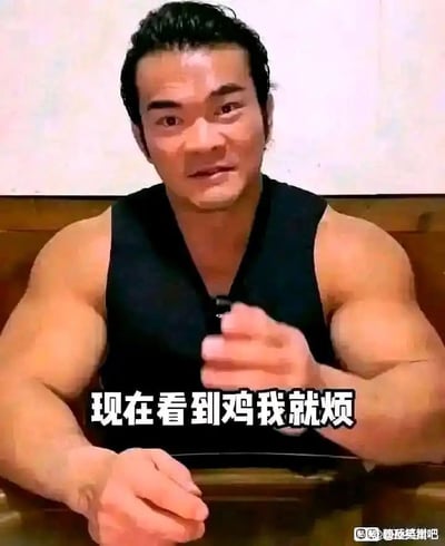 图片中是一位身材非常壮硕的男性，坐在桌前，身穿黑色背心，表现出健身者的典型形象。配文“现在看到鸡我就烦”直译为“见到鸡肉就感到烦躁”，实际是对健身圈常见饮食内容（大量摄入鸡胸肉）进行戏谑。整体表达出针对健身餐单的厌倦与无奈，这也是圈内常见的自嘲，具有强烈的搞笑和苦中作乐的氛围。文本存在谐音和双关：“鸡”不仅代表鸡肉，也有可能涉及网络用语中的“鸡”字的其他引申，增加幽默感。