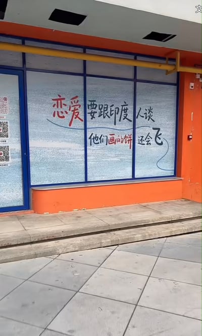 这是一张实拍的街边商铺外观图，商铺外墙为橙色，橱窗边框为蓝色，玻璃贴着磨砂底膜，上面印有手写风格的文字“恋爱要跟印度人谈，他们画的饼还会飞”，搭配简单的曲线装饰，左侧玻璃门处还印有多个二维码，门前是水泥台阶和灰色地砖。内容玩了“画饼”（指口头承诺无法兑现）的网络梗，结合印度飞饼的特色，调侃和印度人恋爱时对方给出的承诺更加虚无缥缈、难以兑现，风格搞笑戏谑。