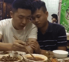 这是一张动态GIF图，呈现了聚餐餐桌场景：一位穿白色短袖的男生正专注地用筷子夹取碗中的食物，身旁穿黑灰条纹短袖的男生亲昵地依偎在他肩头，二人姿态十分亲近。桌上摆放着多道菜肴，整体氛围温馨又带着一丝搞笑感，图中没有双关、谐音梗或暗示内容。