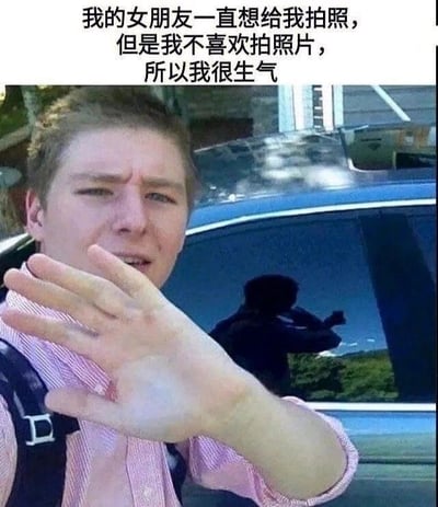 这是一张图文混排的静态单图，画面上方配有文字“我的女朋友一直想给我拍照，但是我不喜欢拍照片，所以我很生气”。图中前景是一位穿着粉色条纹衬衫、背着黑色背包的真实男性，他眉头紧皱，面部表情不悦且带着烦躁，伸出左手做出向前阻挡的动作，似乎在抗拒被拍摄。而在他身后的汽车侧窗玻璃上，清晰地反射出一个举着手机、正在进行拍摄动作的人影，结合上方的文字内容可以推测，这个人影就是他口中想给他拍照的女朋友。这张图的趣味点在于形成了反差：男生表现出强烈的抗拒拍照的情绪，但他此刻的状态已经被成功记录下来，这种矛盾的场景让画面充满了搞笑的氛围，调侃了情侣间一方想记录日常另一方却抗拒的常见互动。