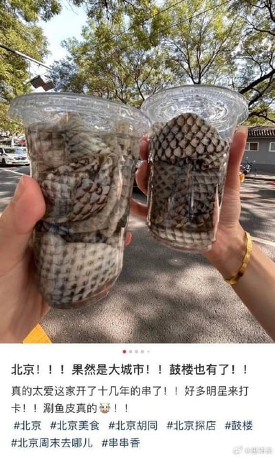 这是一张美食探店分享内容，画面上方是两只手捧着两杯装在透明塑料杯里的涮鱼皮，鱼皮带有清晰的纹理，背景是北京胡同的户外场景，能看到行道树、街道和灰瓦老房，充满老北京氛围。下方是博主的文字分享，激动地推荐这家开了十几年的串串店，提到众多明星前来打卡，对涮鱼皮的口感表示惊叹，并附带多个北京美食、探店相关的话题标签，整体是向网友安利这家位于北京鼓楼的特色街边美食。