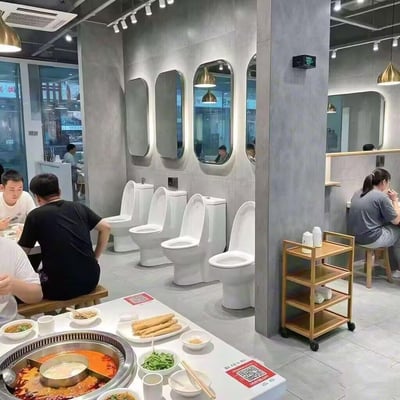 这是一家主打猎奇风格的马桶主题火锅店，店内靠墙区域摆放着多座白色抽水马桶作为特色装饰，与前方的火锅餐桌形成强烈的视觉反差。顾客们坐在简易木凳上，围着餐桌享用火锅，桌上摆放着鸳鸯火锅锅底、绿色蔬菜、炸物等餐品，店铺整体为工业风装修，灰色墙面搭配椭圆形镜面，营造出荒诞又有趣的就餐氛围，没有双关或谐音梗，纯粹以猎奇的马桶装饰打造独特主题。
